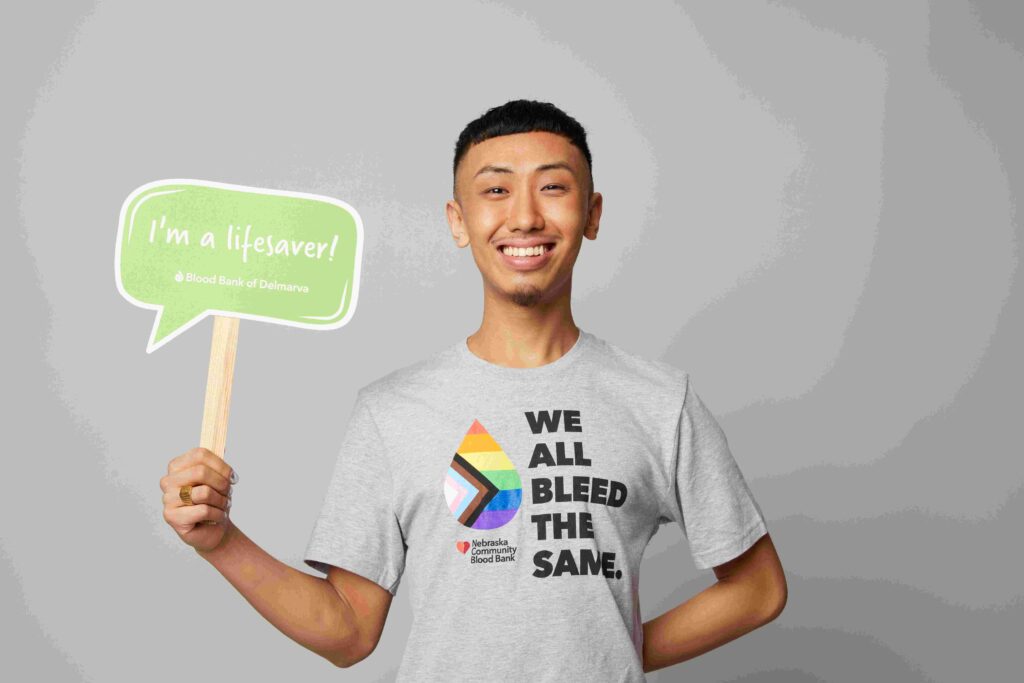 Smiling person wearing a tshirt reading We All Bleed the Same holding a sign that reads I'm a Lifesaver.